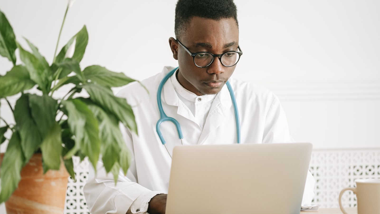 Doctor wearing a stethoscope works on a laptop at a bright office desk.