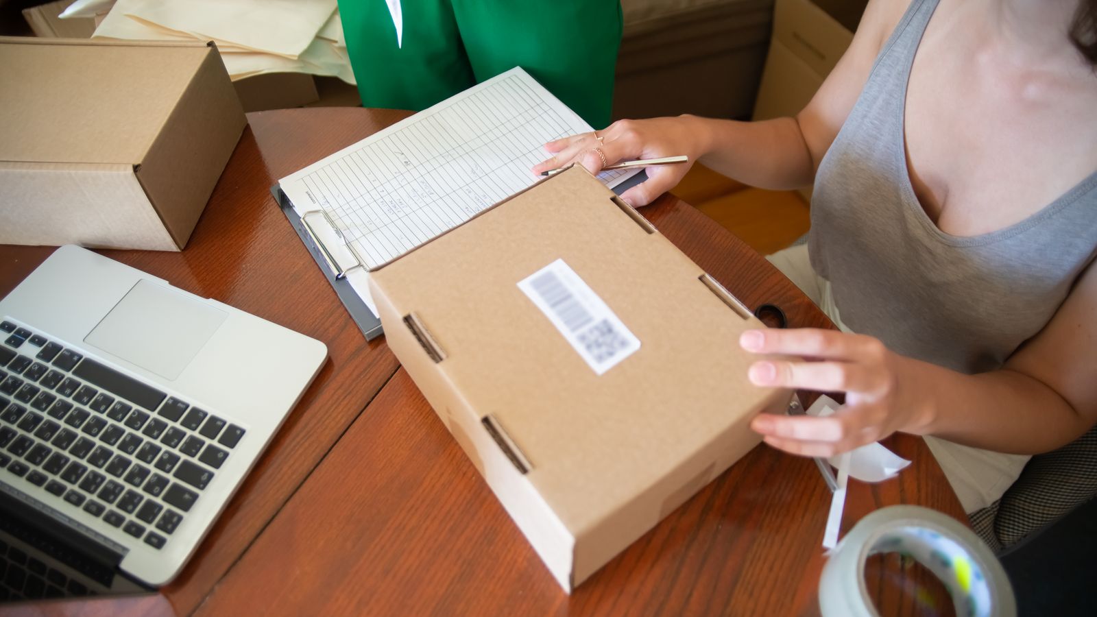 An online seller packs an order beside a laptop and paper order log.
