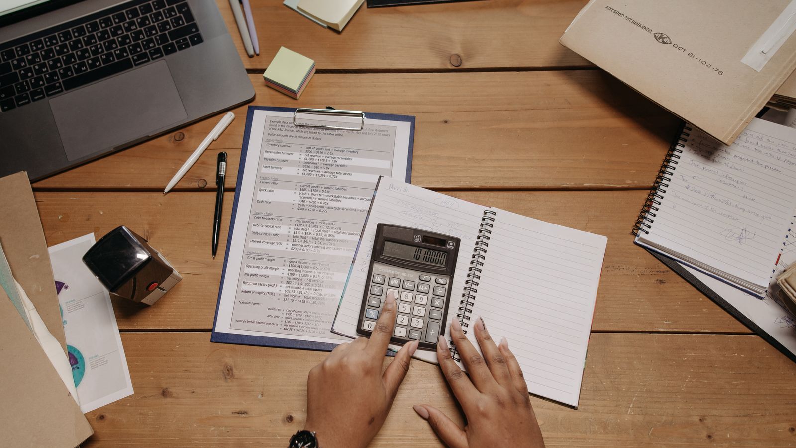 Hands use a calculator beside a laptop, financial worksheets and notebooks.
